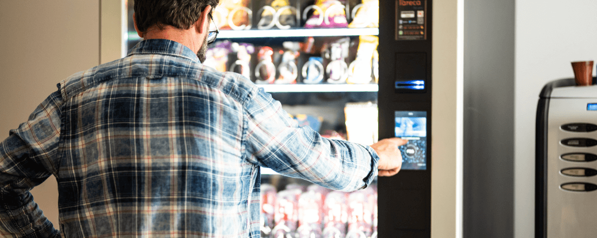 Man selecting a snack from a vending machine, with his finger on the button, wearing a blue square shirt.