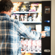 Man selecting a snack from a vending machine, with his finger on the button, wearing a blue square shirt.