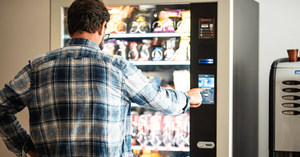 Man selecting a snack from a vending machine, with his finger on the button, wearing a blue square shirt.